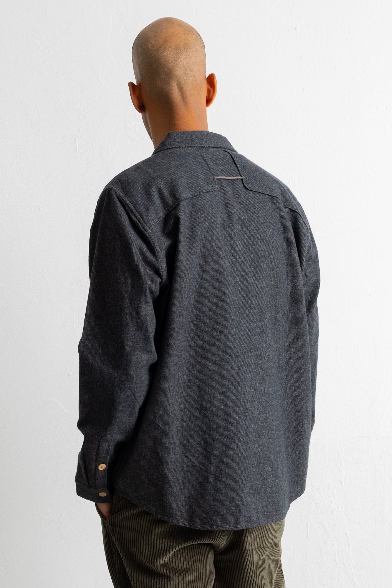 A man viewed from behind wearing a dark charcoal gray long-sleeve linen shirt and brown corduroy trousers, standing against a plain white wall.