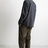 A man standing in profile wearing a dark charcoal gray long-sleeve button-up shirt, olive green corduroy pants, and black canvas sneakers against a plain white studio background.