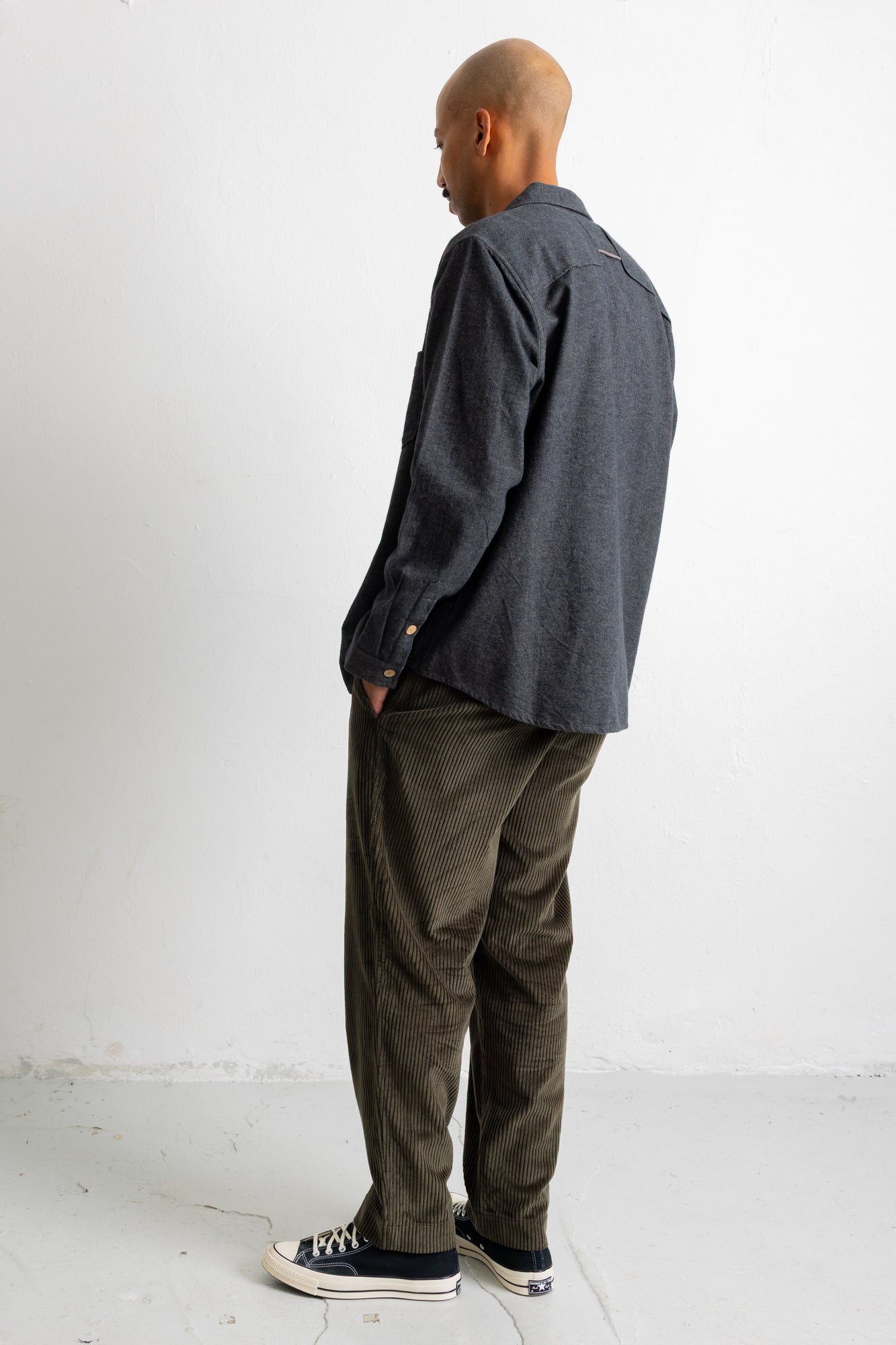 A man standing in profile wearing a dark charcoal gray long-sleeve button-up shirt, olive green corduroy pants, and black canvas sneakers against a plain white studio background.