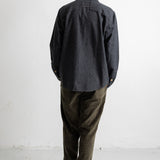 A man viewed from behind wearing a dark charcoal gray long-sleeve button-up shirt, olive green corduroy pants, and black canvas sneakers with white soles, standing against a plain white wall.
