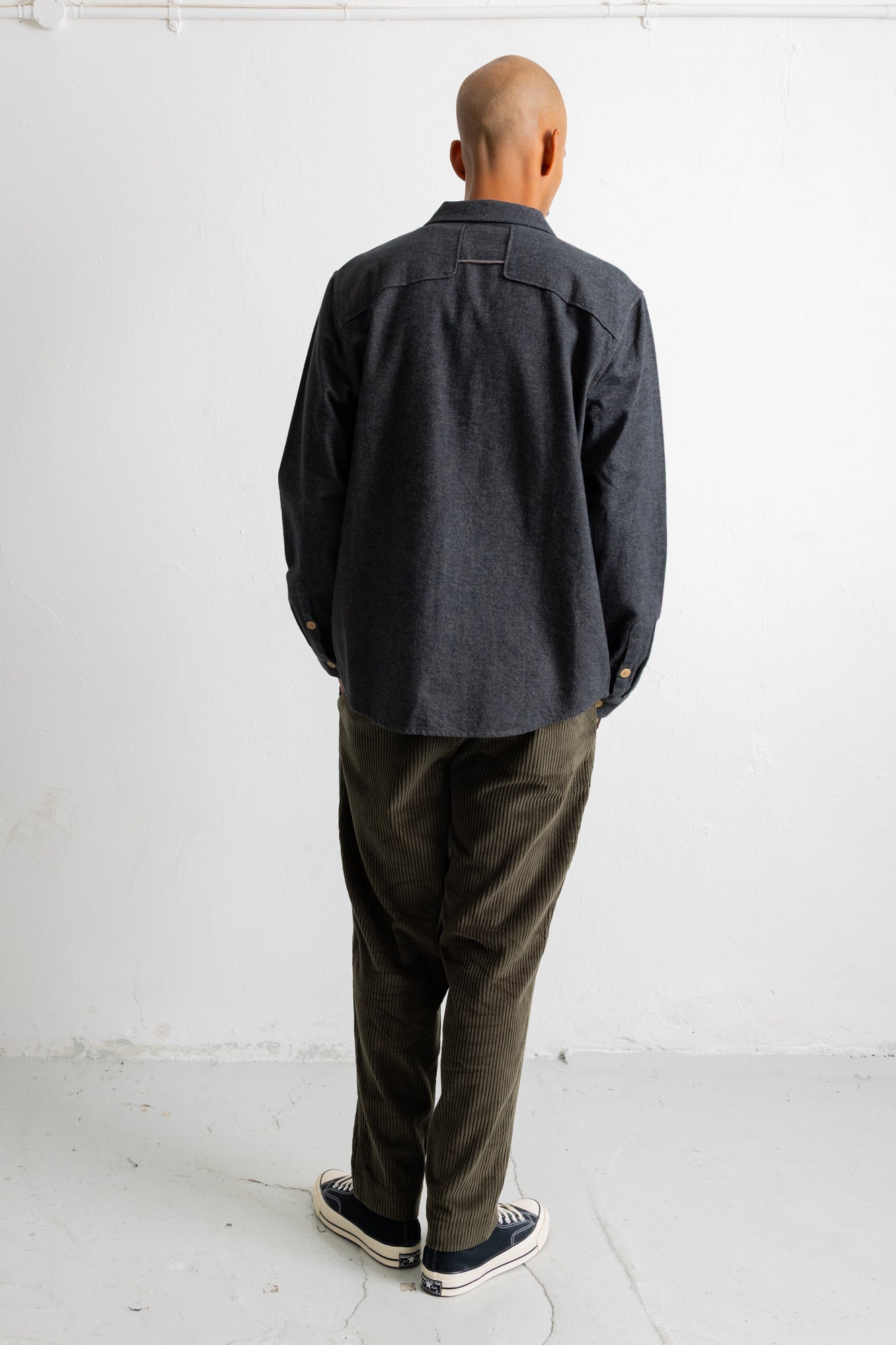 A man viewed from behind wearing a dark charcoal gray long-sleeve button-up shirt, olive green corduroy pants, and black canvas sneakers with white soles, standing against a plain white wall.