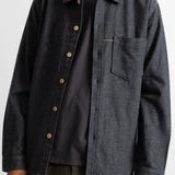 A man wearing a dark charcoal gray button-up overshirt with brown buttons over a black t-shirt and striped pants, shown from the neck down against a light gray background.