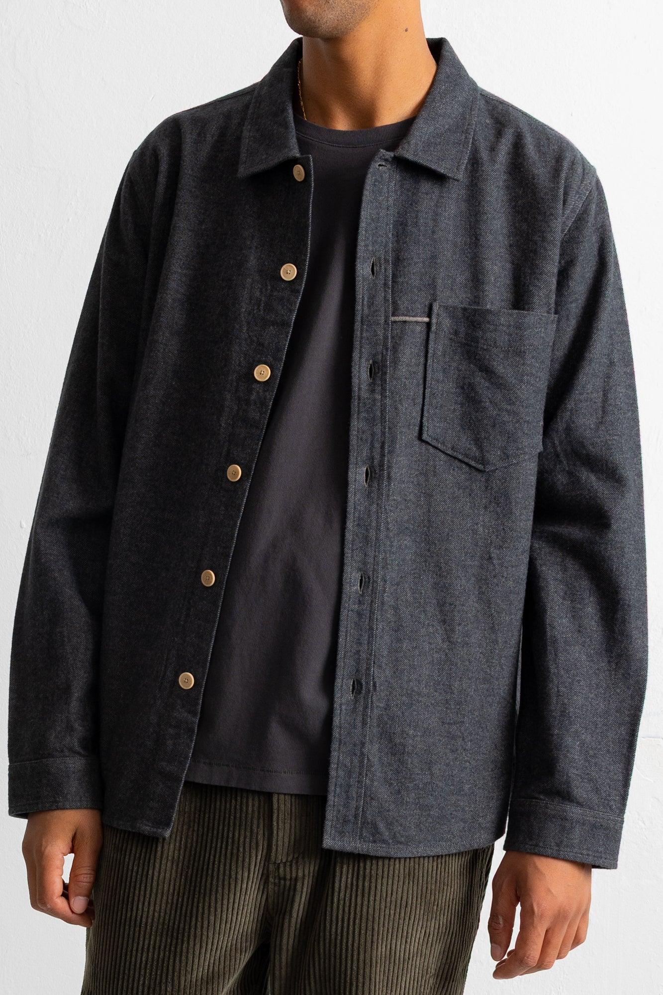 A man wearing a dark charcoal gray button-up overshirt with brown buttons over a black t-shirt and striped pants, shown from the neck down against a light gray background.