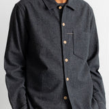 A man wearing a dark charcoal gray long-sleeve button-up shirt with a chest pocket, paired with brown corduroy trousers, against a plain white background.