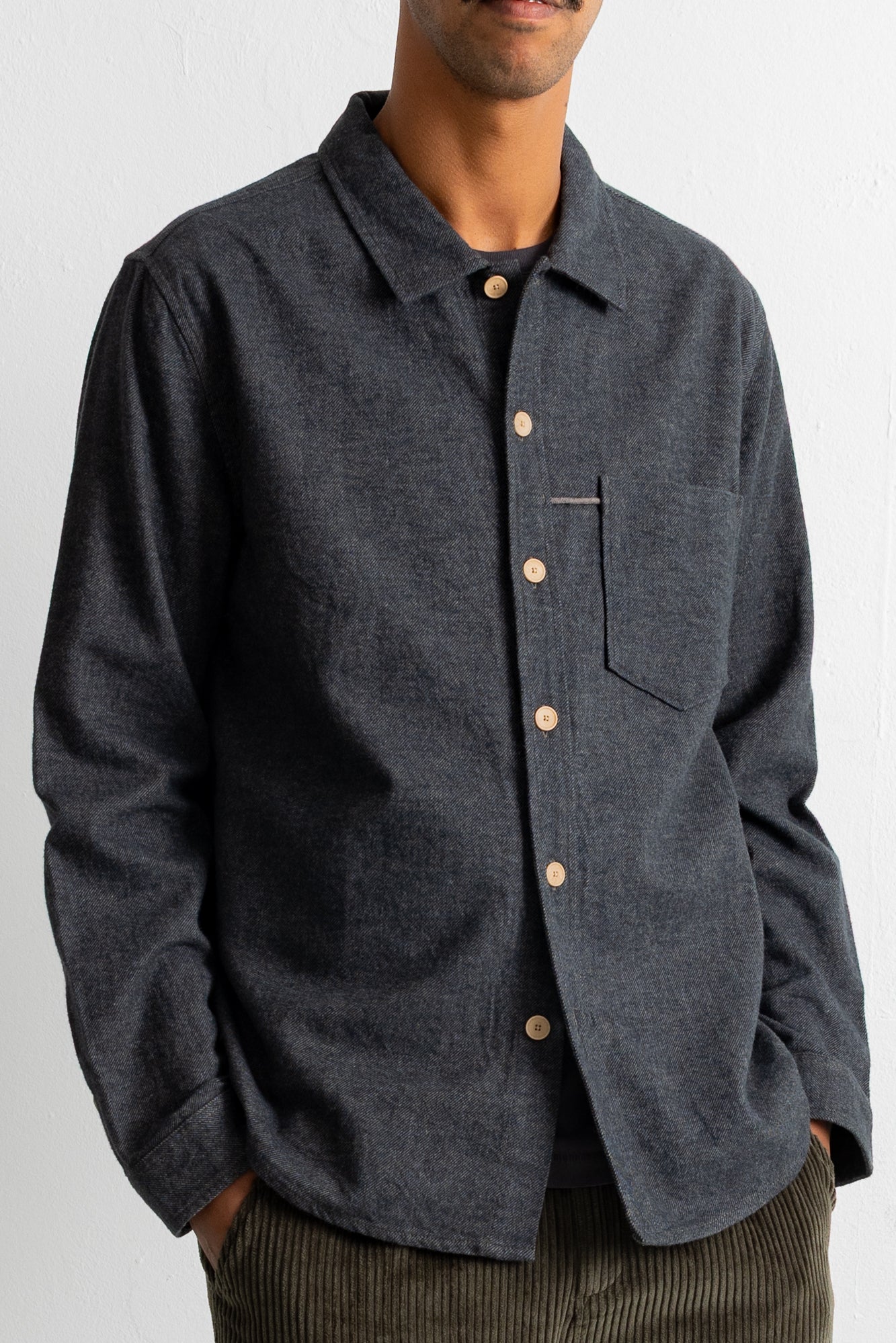 A man wearing a dark charcoal gray long-sleeve button-up shirt with a chest pocket, paired with brown corduroy trousers, against a plain white background.