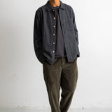 A man wearing a dark charcoal gray button-up overshirt, olive green corduroy pants, and black-and-white sneakers, standing against a plain white wall.