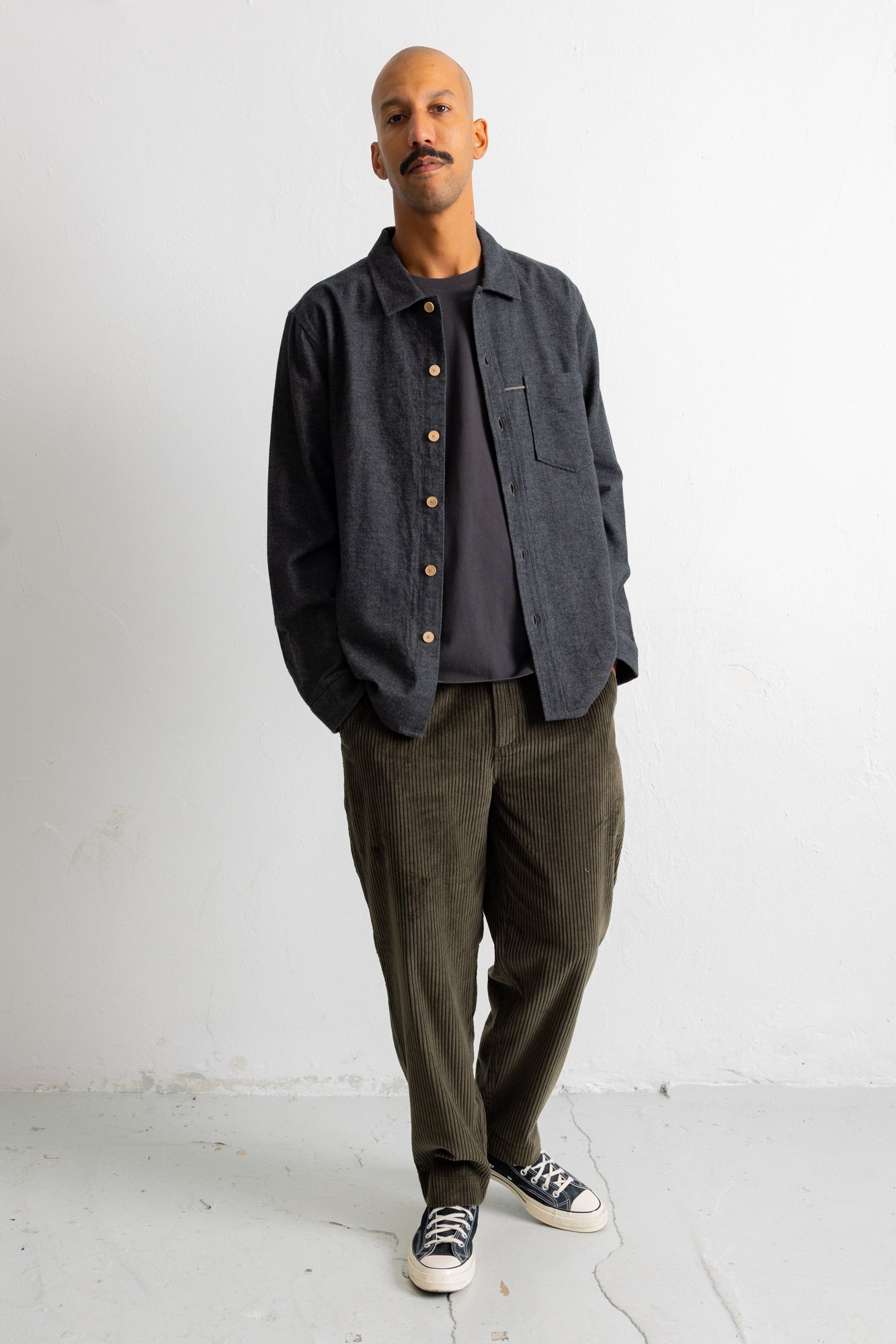A man wearing a dark charcoal gray button-up overshirt, olive green corduroy pants, and black-and-white sneakers, standing against a plain white wall.