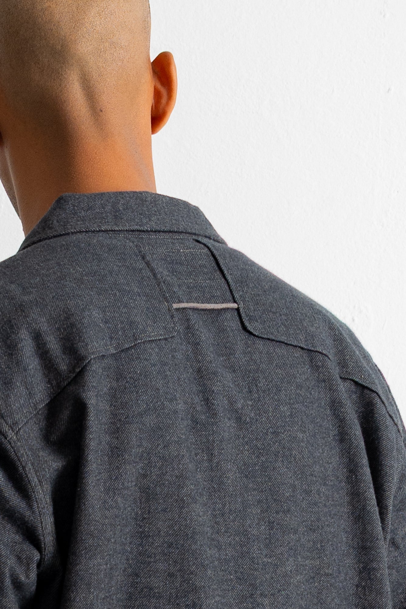 Close-up back view of a person wearing a dark charcoal gray textured overshirt or shirt jacket against a plain white wall.