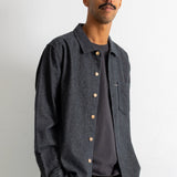 A man wearing a dark charcoal gray long-sleeve button-up shirt over a dark gray t-shirt, paired with brown corduroy trousers, standing against a plain white wall.