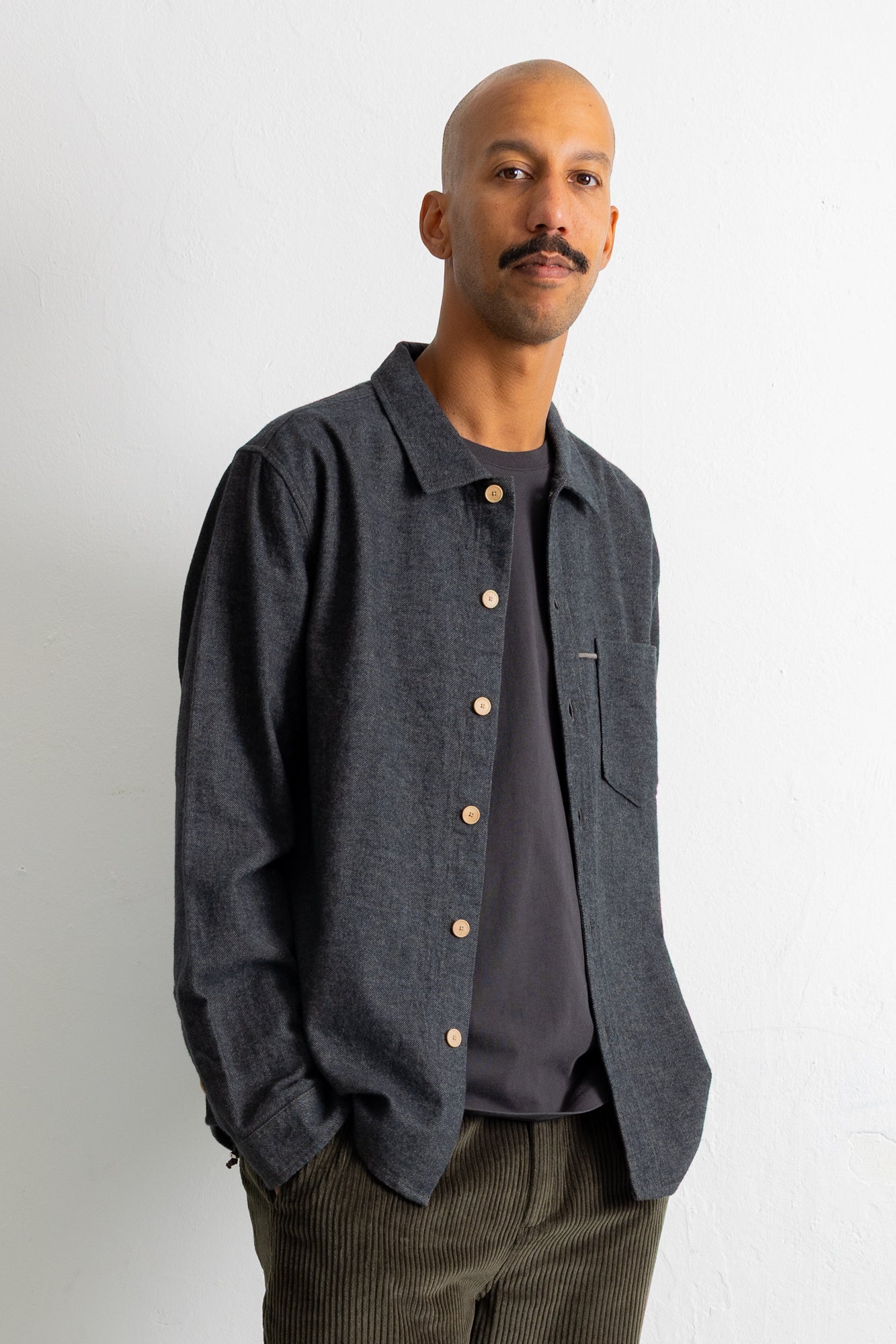 A man wearing a dark charcoal gray long-sleeve button-up shirt over a dark gray t-shirt, paired with brown corduroy trousers, standing against a plain white wall.