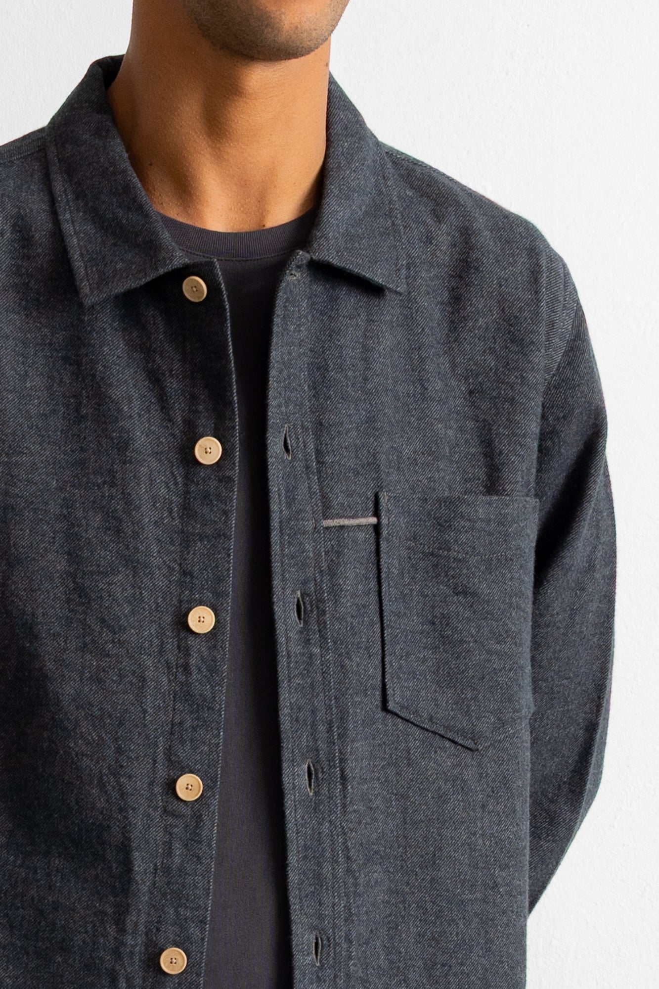 A close-up of a person wearing a dark charcoal gray textured overshirt with light brown buttons over a black t-shirt, against a plain white background.