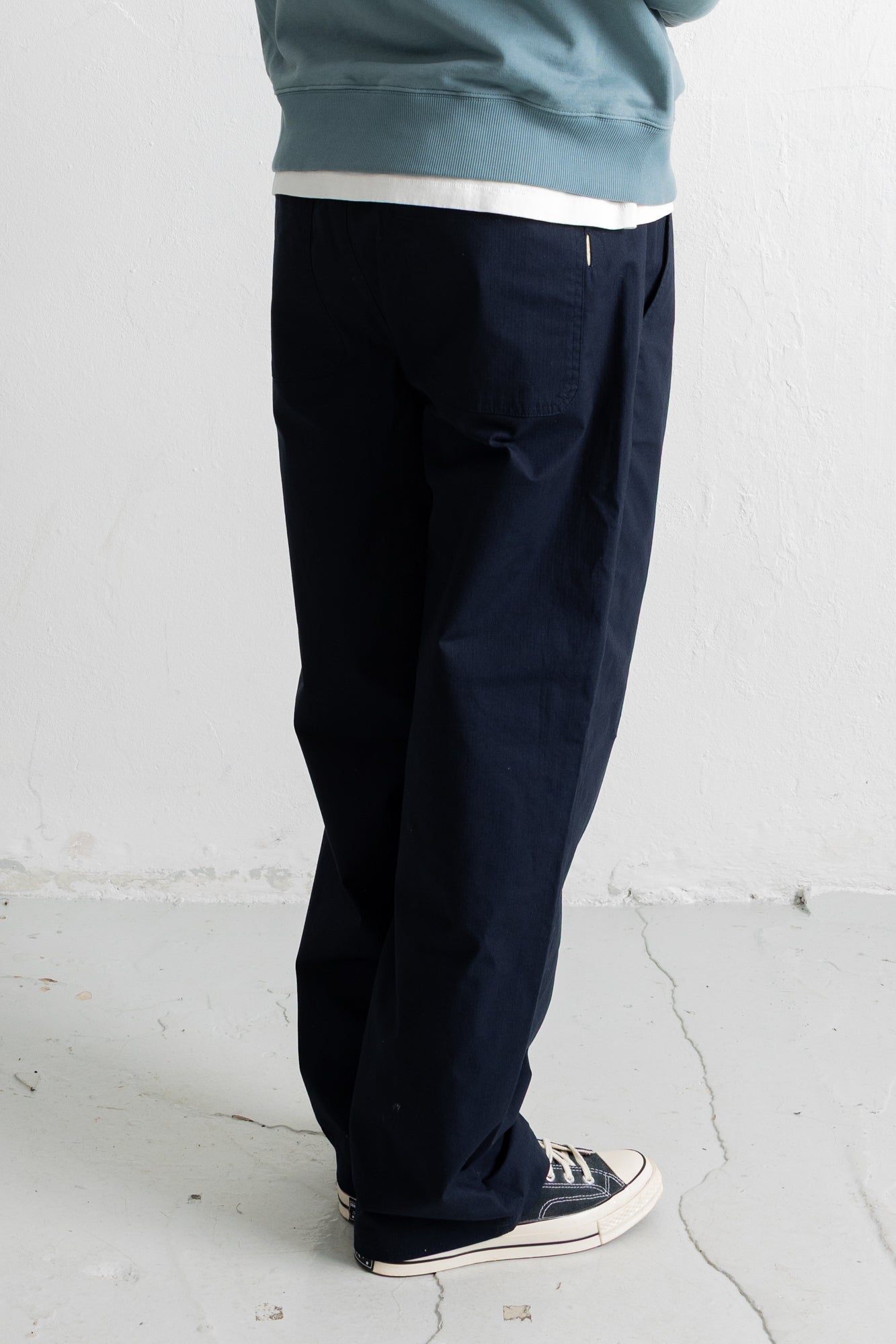 A person shown from the waist down wearing dark navy blue relaxed-fit trousers, a light blue sweatshirt, and navy canvas sneakers with white soles, standing against a white wall.