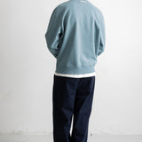 A man viewed from behind wearing a dusty blue crewneck sweatshirt, dark navy pants, and white sneakers, standing against a plain white wall.