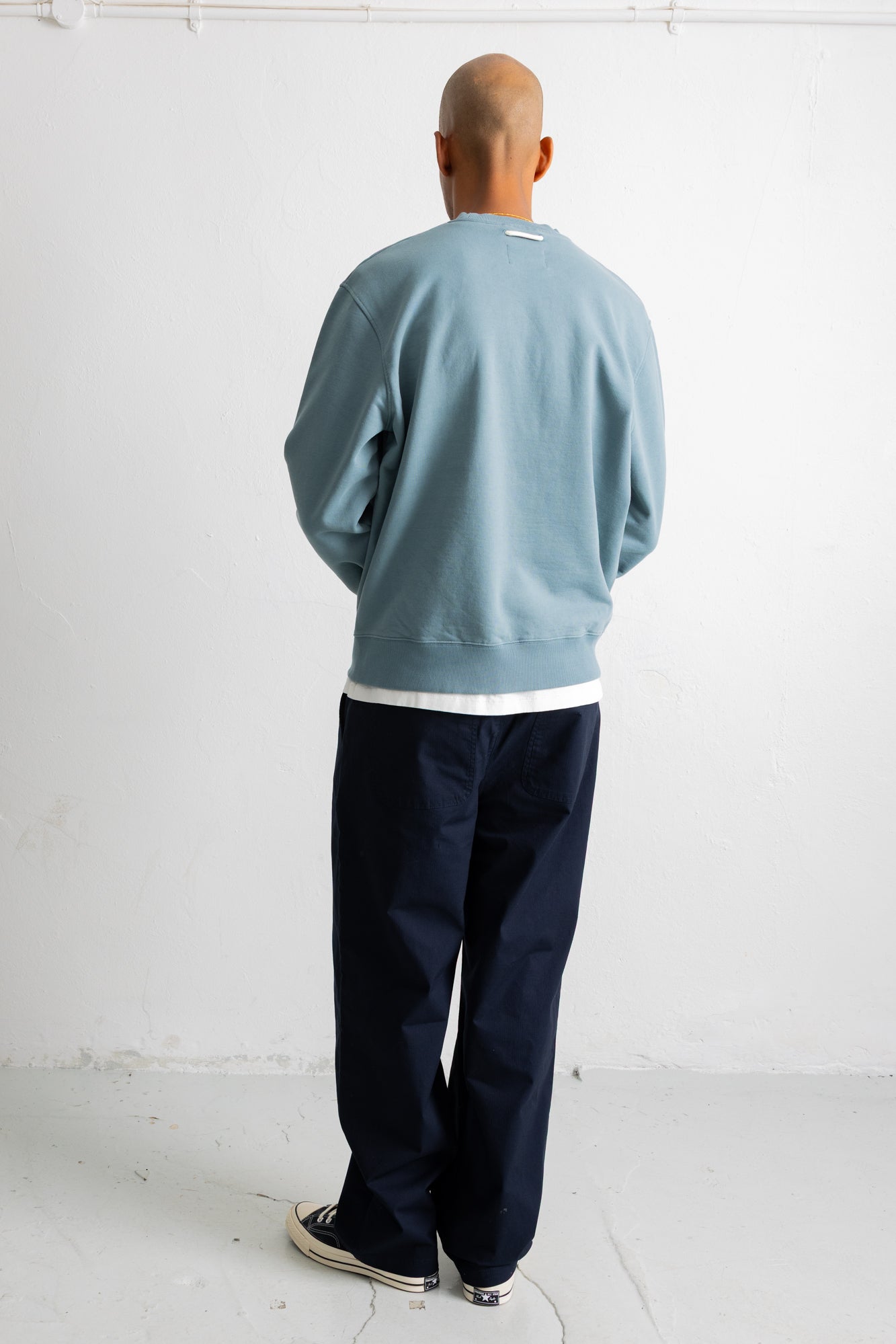 A man viewed from behind wearing a dusty blue crewneck sweatshirt, dark navy pants, and white sneakers, standing against a plain white wall.