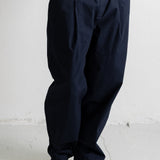 A person wearing dark navy pleated trousers, a light blue sweatshirt, and navy canvas sneakers with cream soles, standing against a light gray wall.