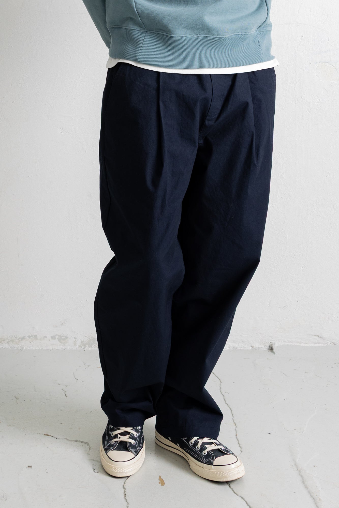A person wearing dark navy pleated trousers, a light blue sweatshirt, and navy canvas sneakers with cream soles, standing against a light gray wall.