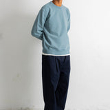 A man wearing a light blue crewneck sweatshirt, dark navy pants, and black canvas sneakers, standing against a white wall.