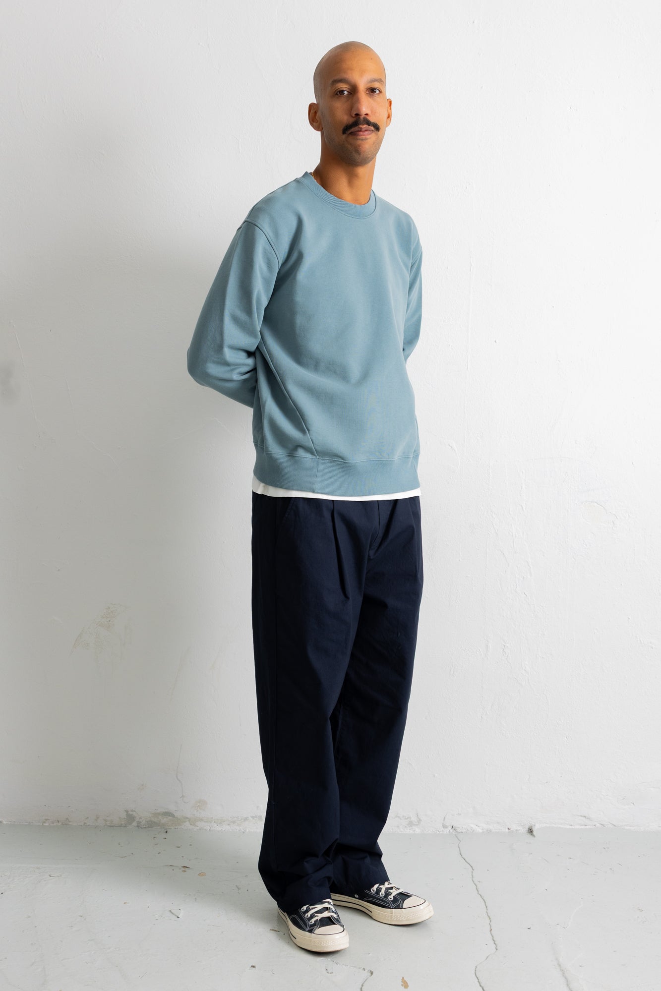 A man wearing a light blue crewneck sweatshirt, dark navy pants, and black canvas sneakers, standing against a white wall.