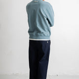 A man viewed from behind wearing a dusty blue crewneck sweatshirt, dark navy pants, and white sneakers, standing against a white wall.