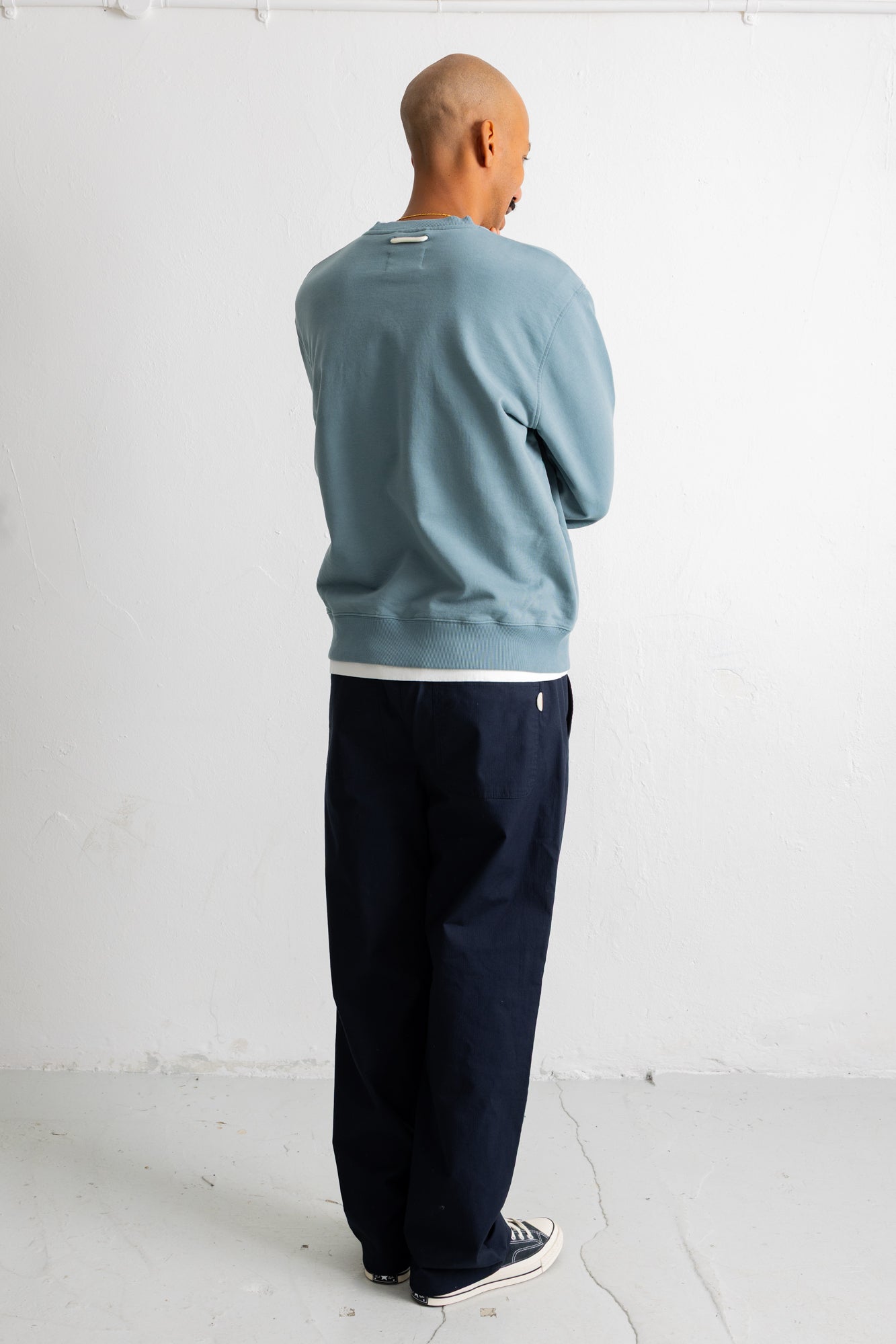 A man viewed from behind wearing a dusty blue crewneck sweatshirt, dark navy pants, and white sneakers, standing against a white wall.