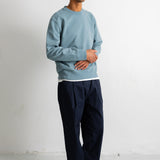 A man wearing a light blue crewneck sweatshirt, dark navy pleated trousers, and black canvas sneakers, standing against a white studio wall.