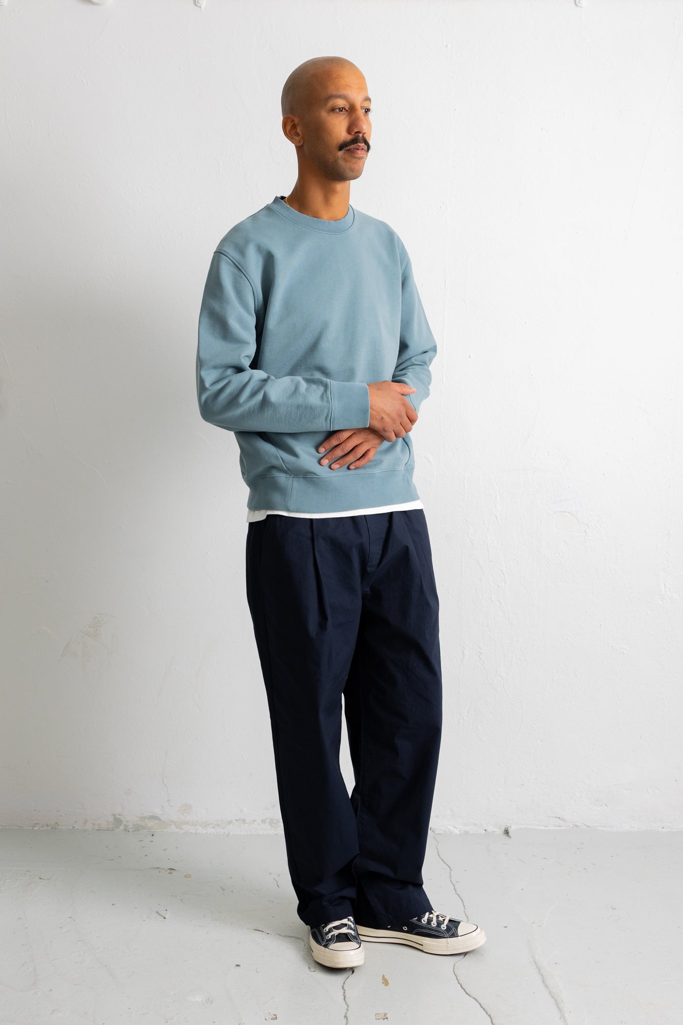 A man wearing a light blue crewneck sweatshirt, dark navy pleated trousers, and black canvas sneakers, standing against a white studio wall.