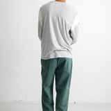 A man viewed from behind wearing a light gray long-sleeve shirt and dark green trousers, standing against a plain white wall.