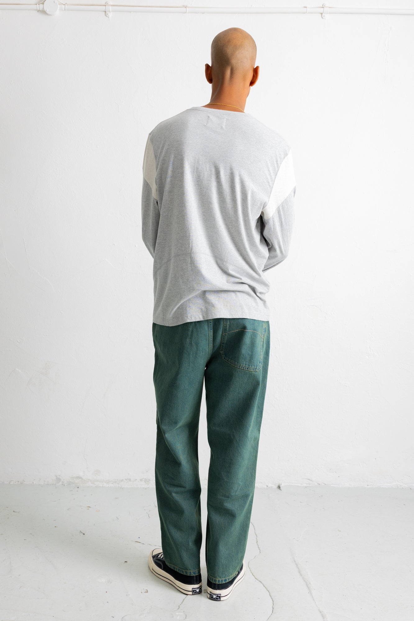 A man viewed from behind wearing a light gray long-sleeve shirt and dark green trousers, standing against a plain white wall.