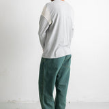A man shown from the back wearing a light gray long-sleeve shirt with white shoulder panels and dark green corduroy pants, standing against a white wall.