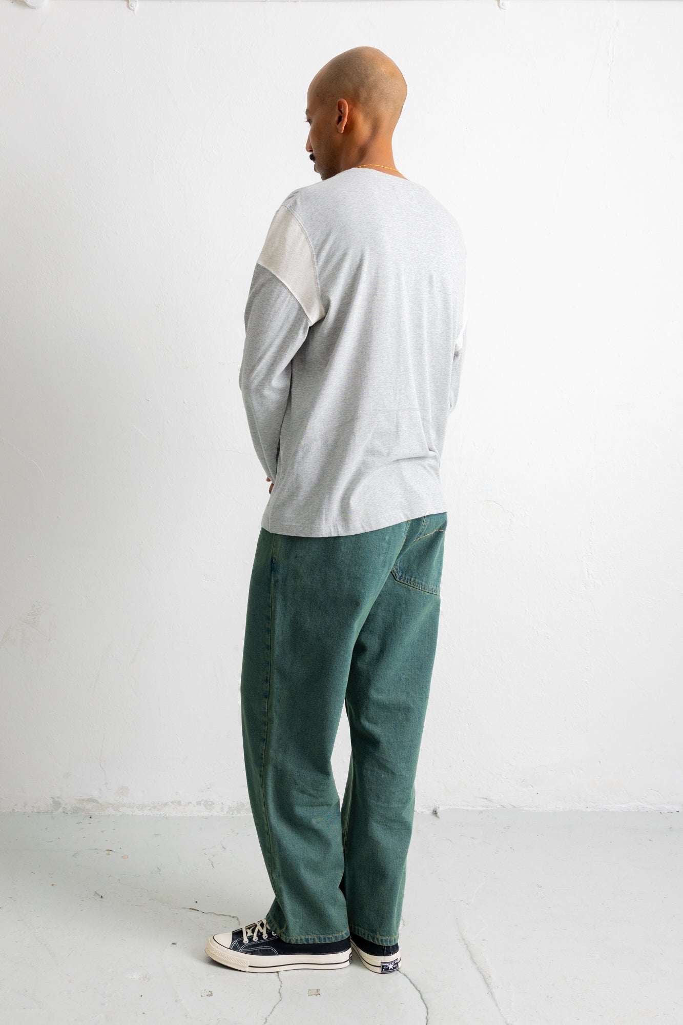 A man shown from the back wearing a light gray long-sleeve shirt with white shoulder panels and dark green corduroy pants, standing against a white wall.