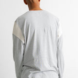 A man viewed from behind wearing a light gray long-sleeve shirt with contrasting cream panels on the upper sleeves, paired with teal jeans, against a plain white background.