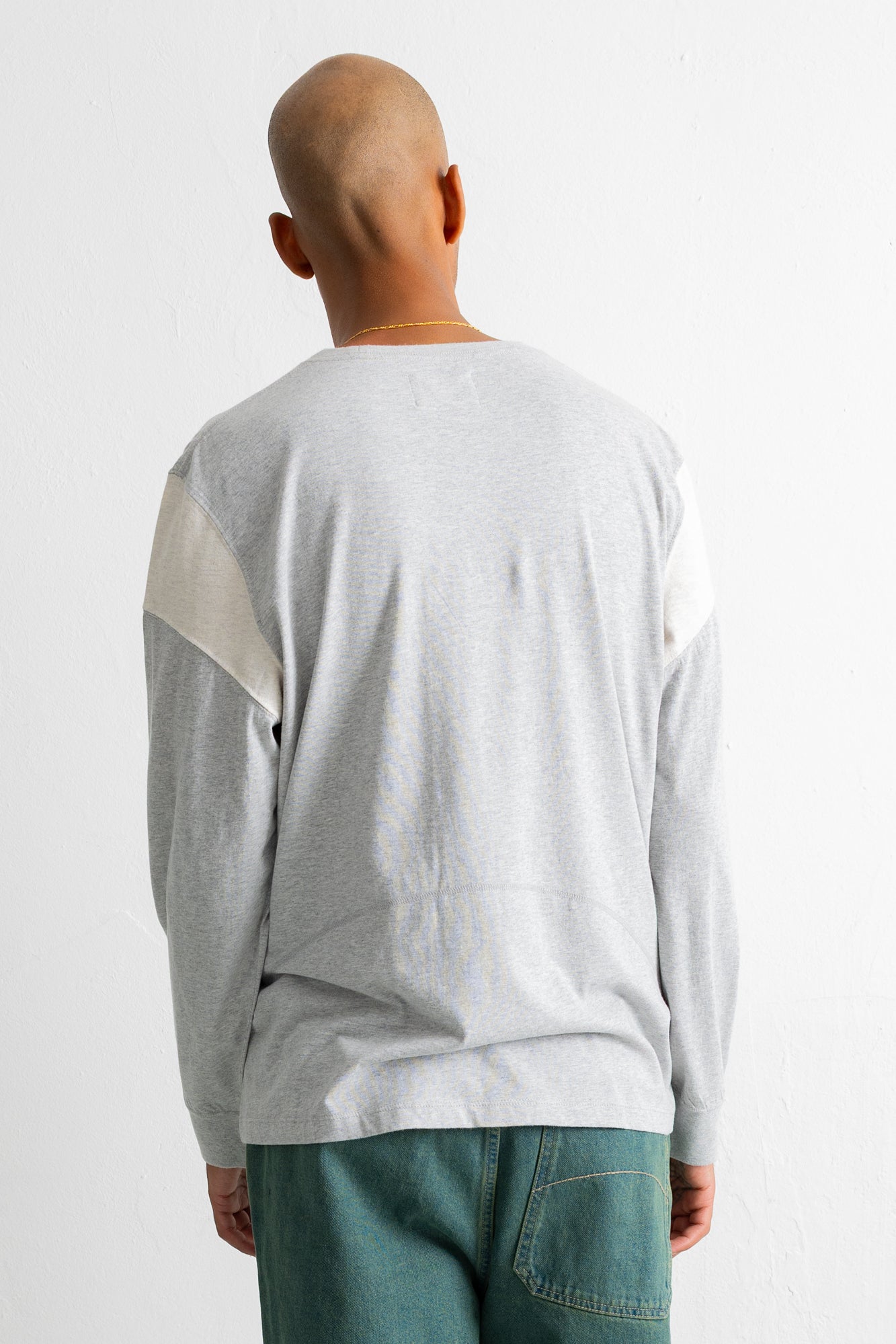 A man viewed from behind wearing a light gray long-sleeve shirt with contrasting cream panels on the upper sleeves, paired with teal jeans, against a plain white background.