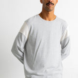 A man wearing a light gray long-sleeve crewneck shirt with contrasting white shoulder panels, paired with teal jeans, photographed against a plain light gray background.