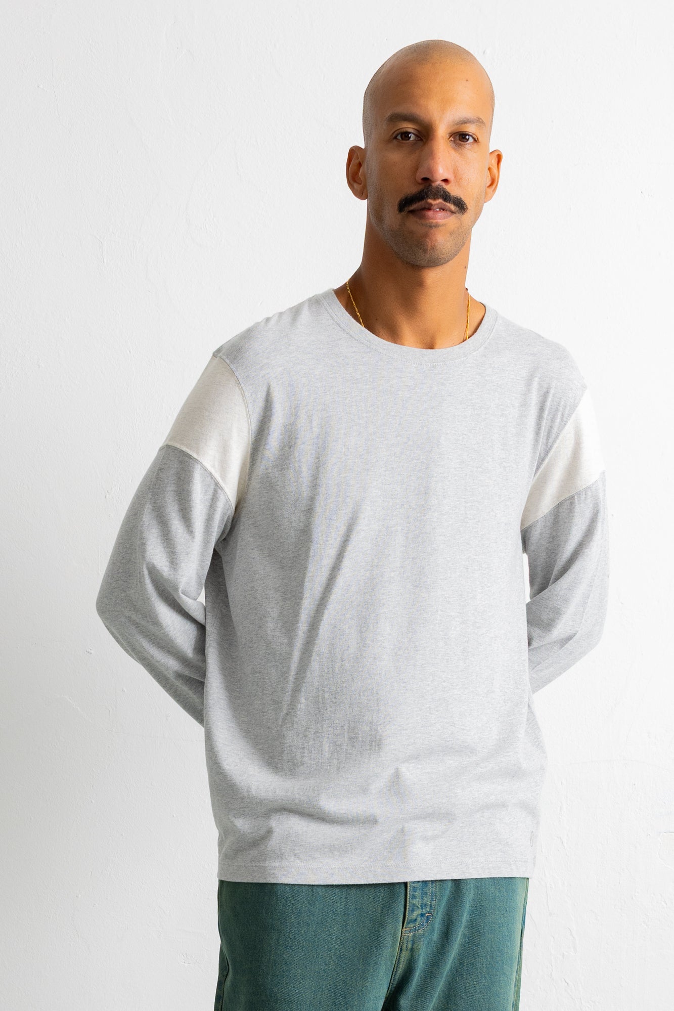 A man wearing a light gray long-sleeve crewneck shirt with contrasting white shoulder panels, paired with teal jeans, photographed against a plain light gray background.
