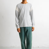 A man wearing a light gray long-sleeve crewneck shirt with contrasting white shoulder panels, paired with green pants and black-and-white sneakers, standing against a white wall.