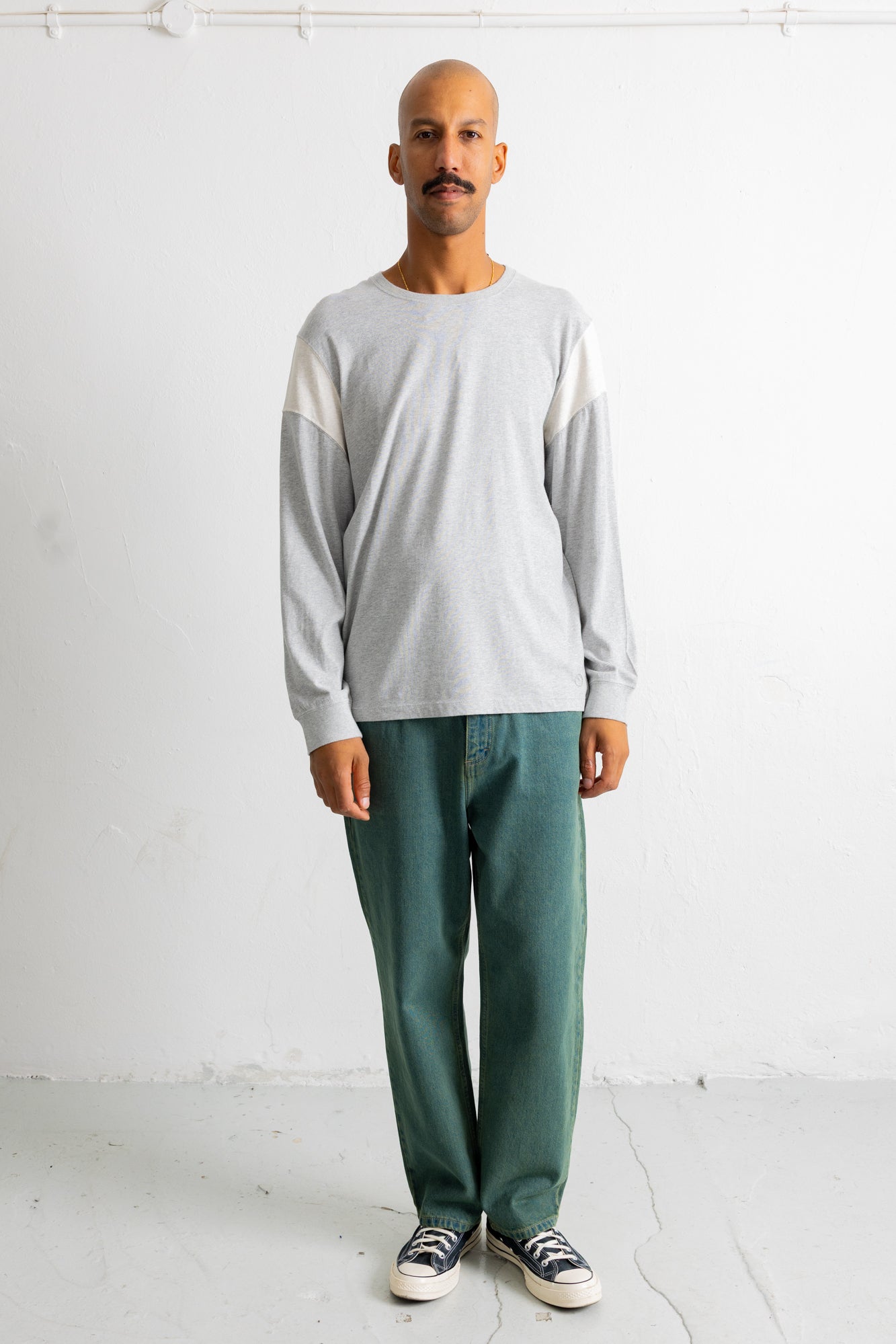 A man wearing a light gray long-sleeve crewneck shirt with contrasting white shoulder panels, paired with green pants and black-and-white sneakers, standing against a white wall.