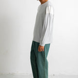 A man stands in profile wearing a light gray long-sleeve top with white shoulder panels, dark green wide-leg pants, and black canvas sneakers against a plain white studio wall.