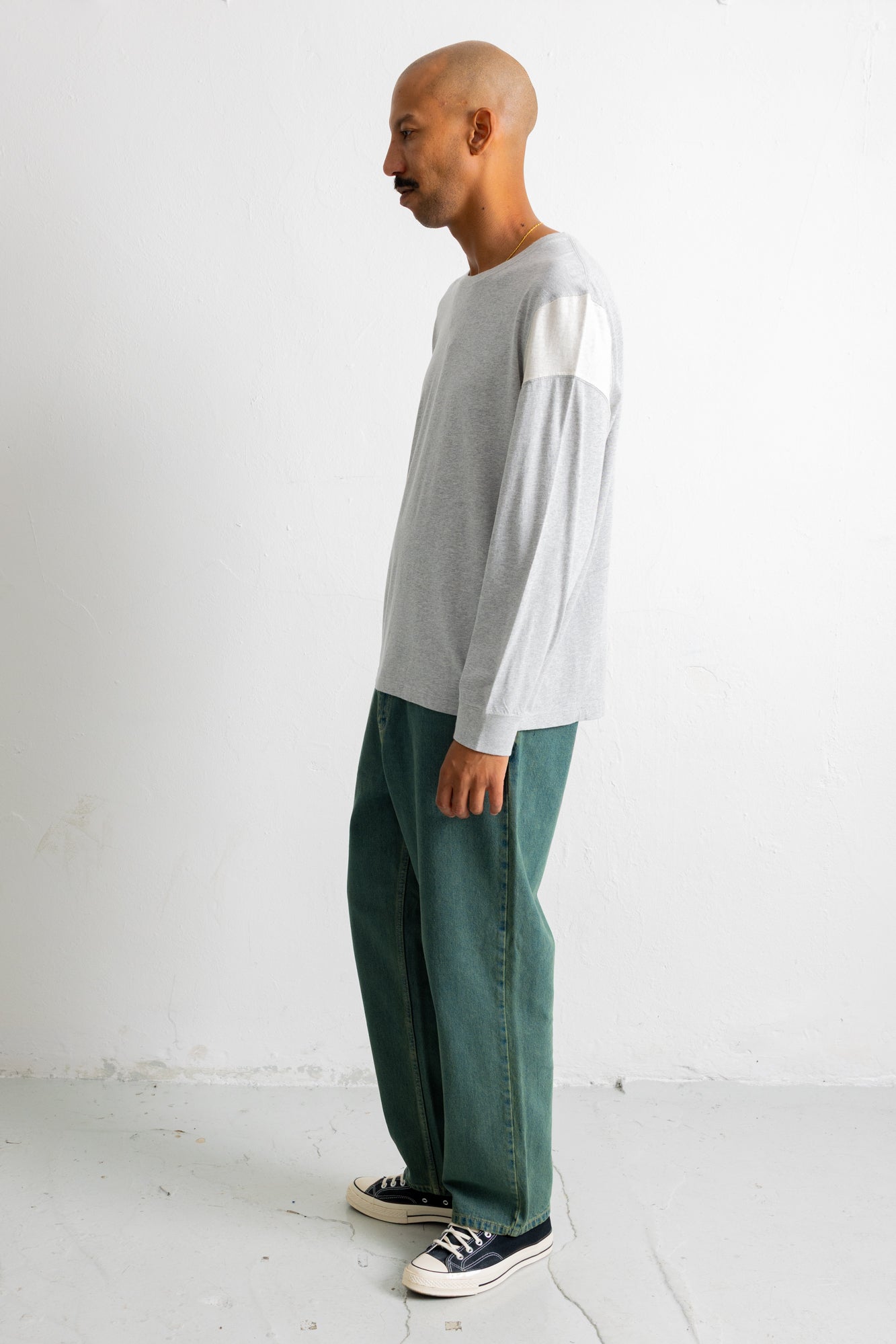 A man stands in profile wearing a light gray long-sleeve top with white shoulder panels, dark green wide-leg pants, and black canvas sneakers against a plain white studio wall.