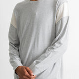 A man wearing a light gray long-sleeve crewneck shirt with contrasting off-white color-blocked shoulder panels, paired with teal jeans against a plain white background.