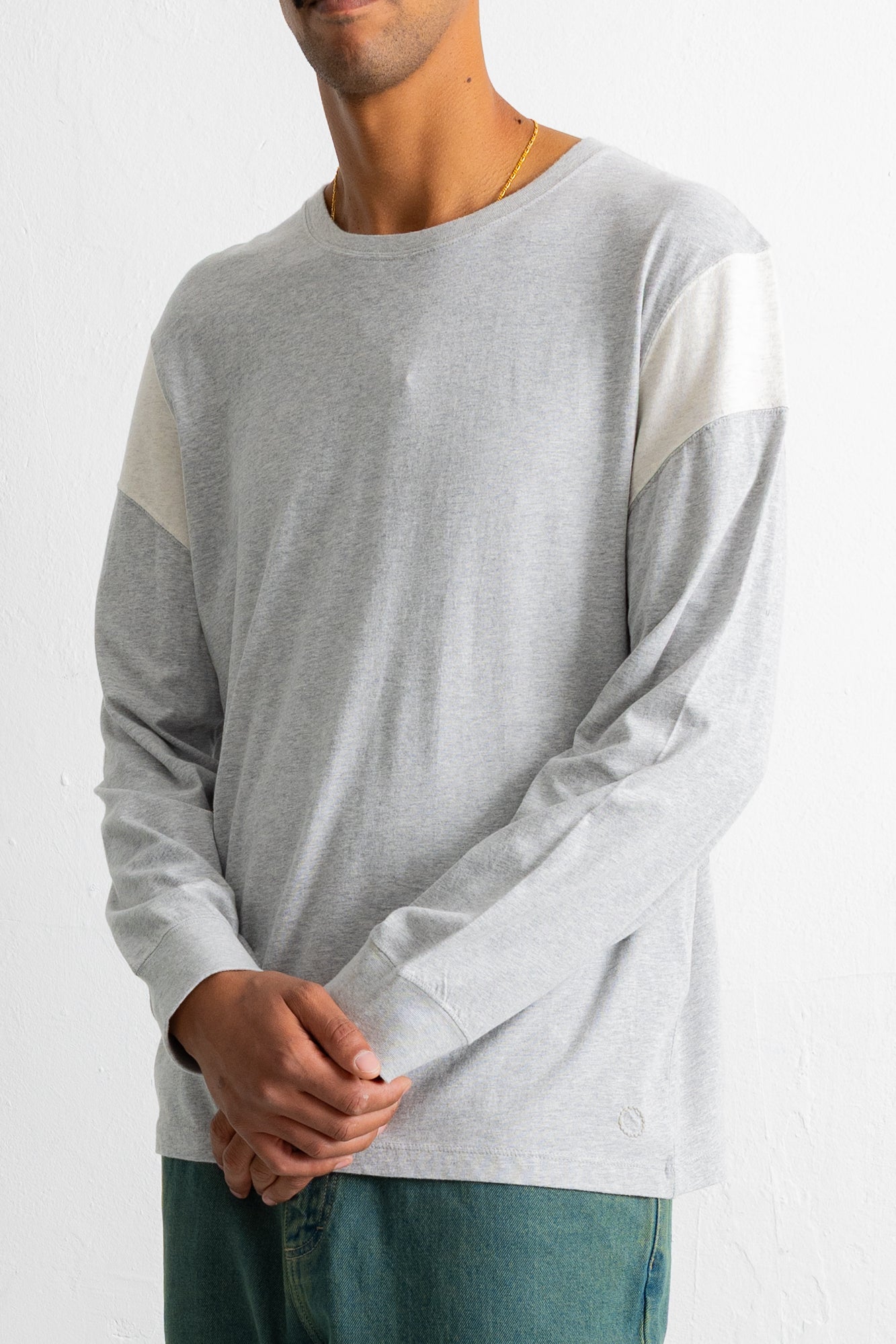 A man wearing a light gray long-sleeve crewneck shirt with contrasting off-white color-blocked shoulder panels, paired with teal jeans against a plain white background.