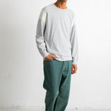 A man wearing a light gray long-sleeve crewneck shirt with contrasting light yellow shoulder panels, paired with dark green relaxed-fit jeans and black canvas sneakers, standing against a white studio wall.