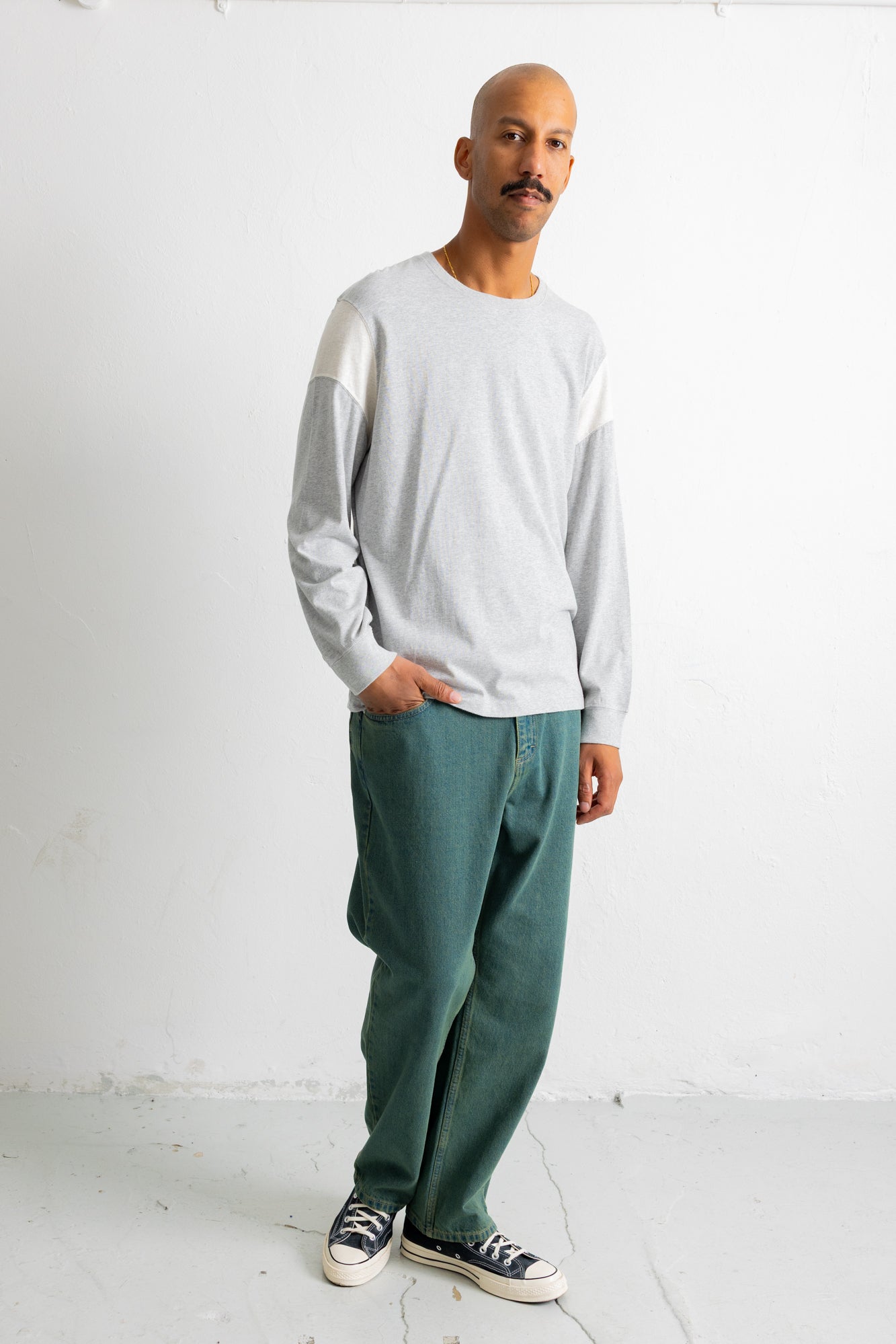 A man wearing a light gray long-sleeve crewneck shirt with contrasting light yellow shoulder panels, paired with dark green relaxed-fit jeans and black canvas sneakers, standing against a white studio wall.