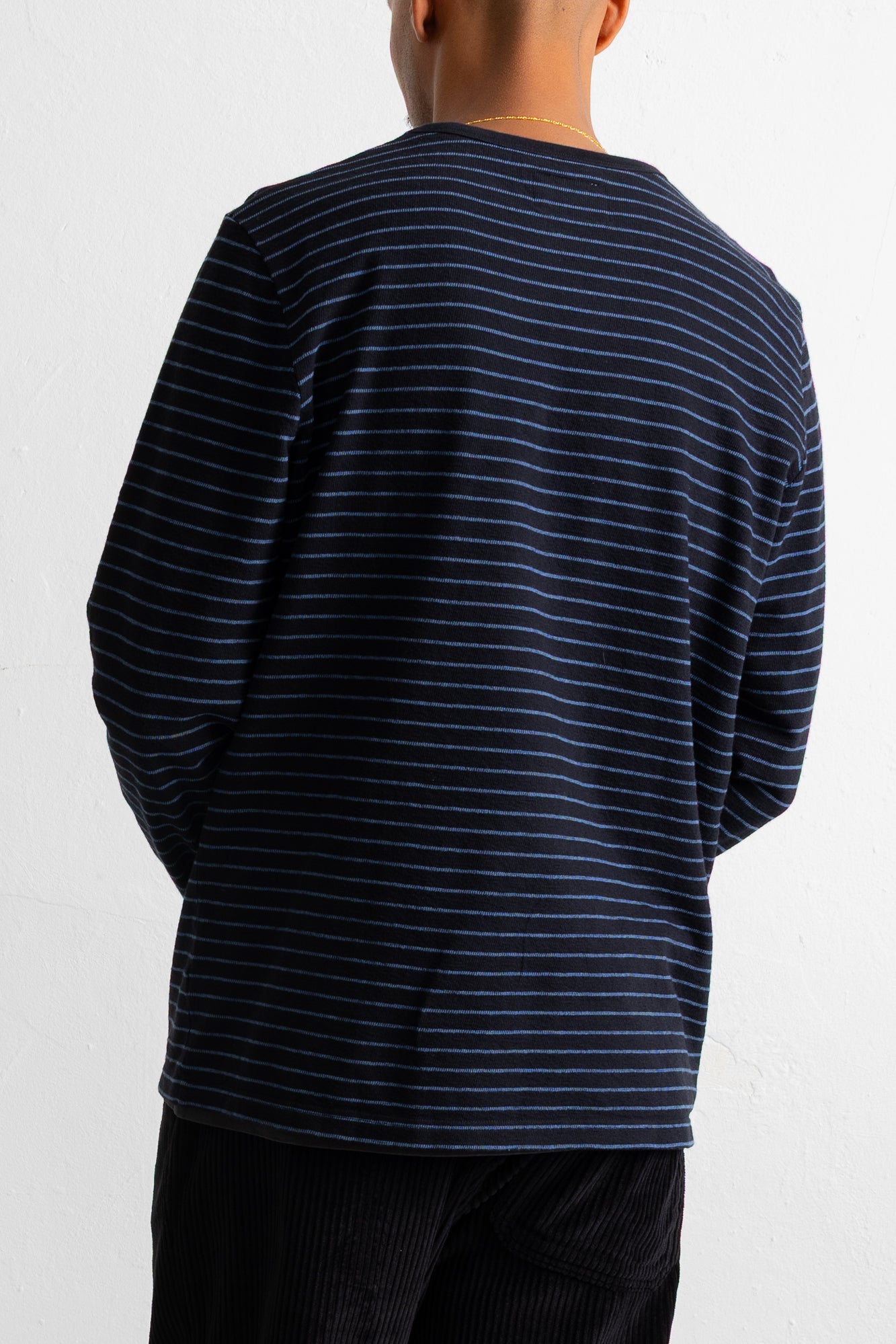 A person shown from the back wearing a navy long-sleeve shirt with thin white horizontal stripes and black corduroy pants against a plain white wall.