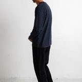 A man in profile wearing a navy blue long-sleeve shirt with thin horizontal dark stripes, black pants, and black canvas sneakers, standing against a plain white wall.
