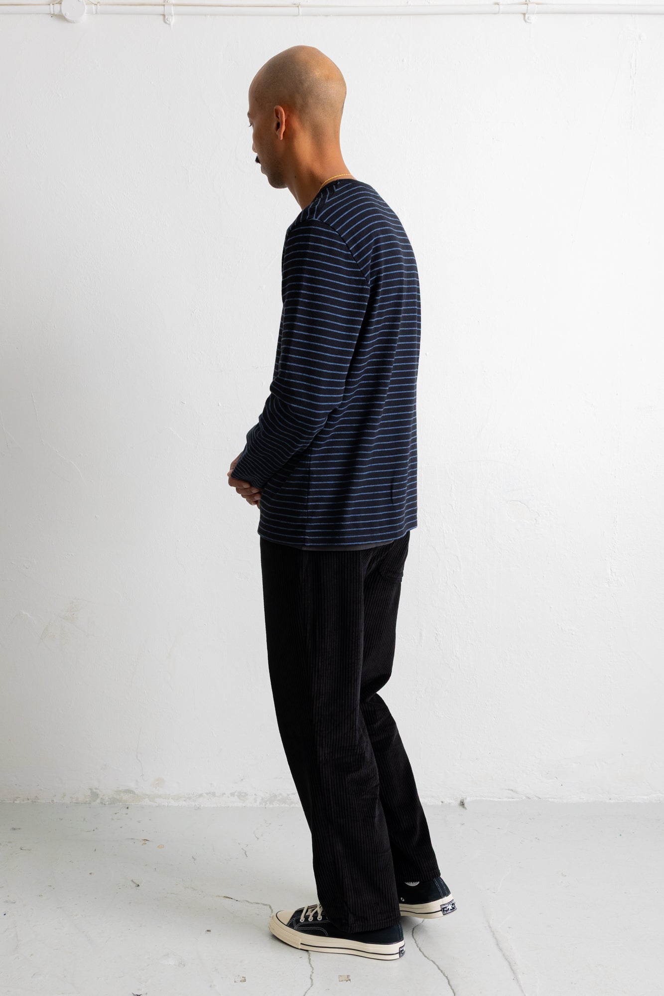 A man in profile wearing a navy blue long-sleeve shirt with thin horizontal dark stripes, black pants, and black canvas sneakers, standing against a plain white wall.