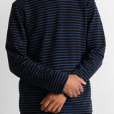 A man wearing a navy blue long-sleeve crewneck shirt with thin white horizontal stripes, paired with black corduroy pants, against a plain white wall.