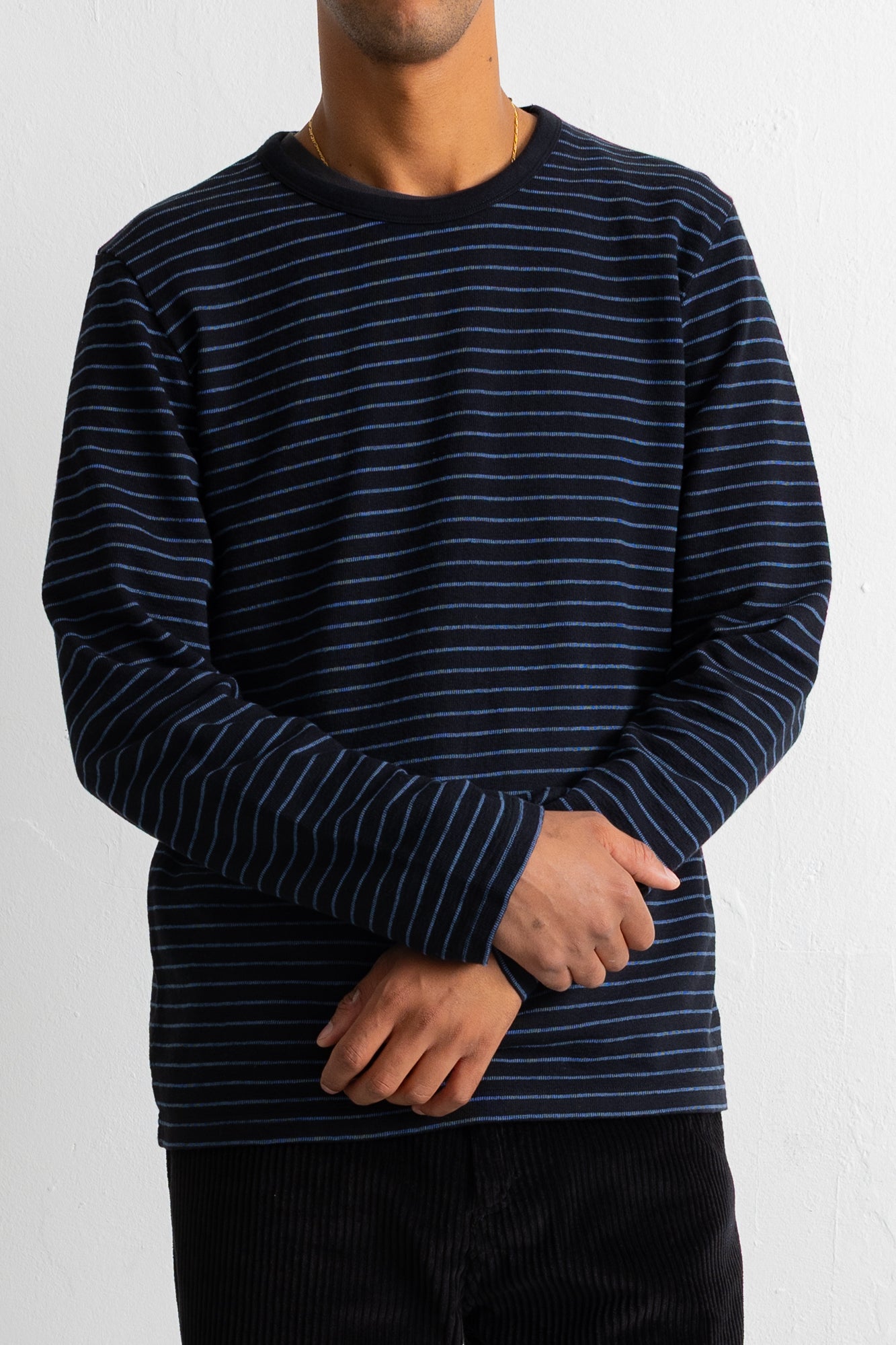 A man wearing a navy blue long-sleeve crewneck shirt with thin white horizontal stripes, paired with black corduroy pants, against a plain white wall.