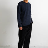 A man wearing a navy blue and black horizontally striped long-sleeve shirt, black trousers, and black canvas sneakers, standing against a plain white studio wall.