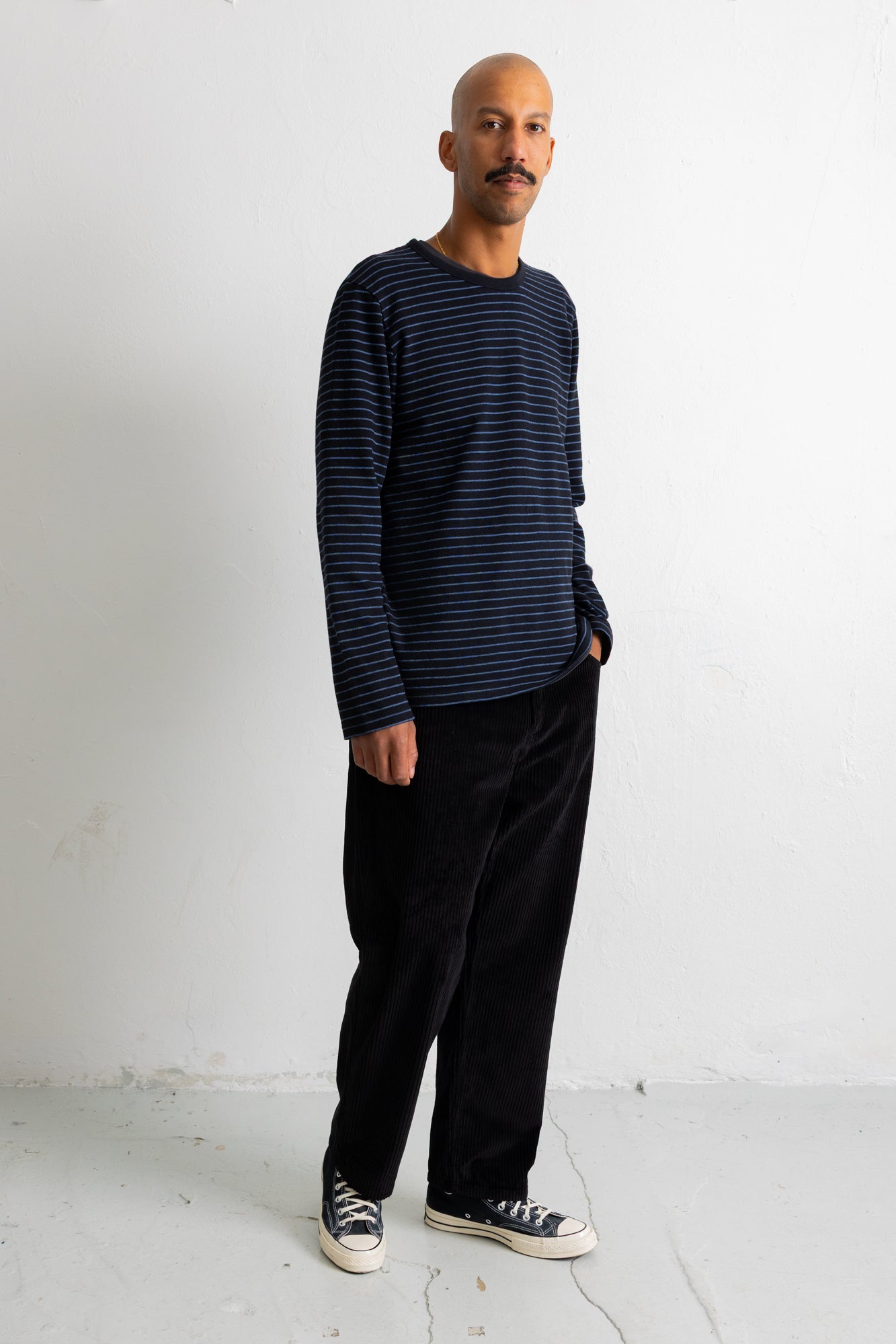 A man wearing a navy blue and black horizontally striped long-sleeve shirt, black trousers, and black canvas sneakers, standing against a plain white studio wall.