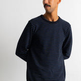 A man wearing a navy blue long-sleeve shirt with thin white horizontal stripes, standing against a plain light gray wall.